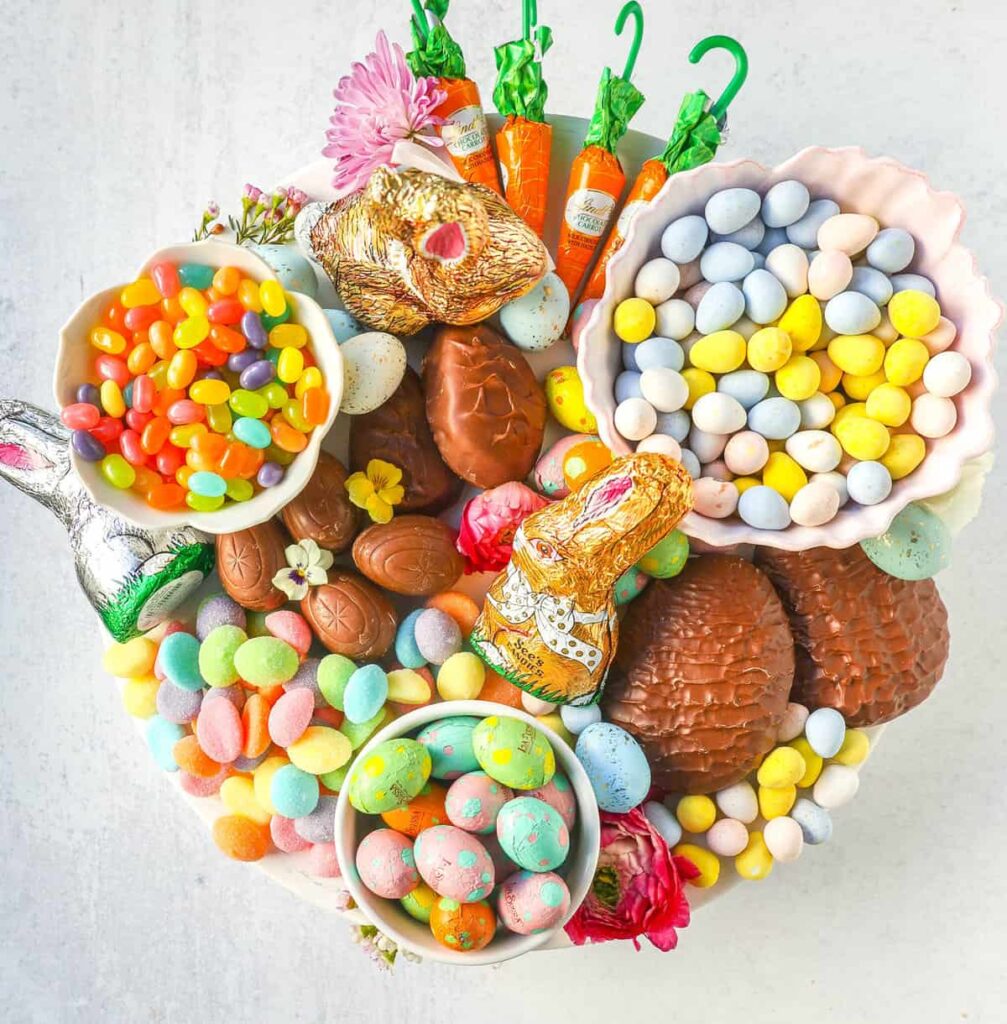 A round white plate overflowing with Easter sweets including foil-wrapped chocolate bunnies, large chocolate eggs, and several bowls of jelly beans and pastel malt balls. Small pink and yellow flowers are tucked between the candies, alongside orange foil-wrapped chocolate carrots.