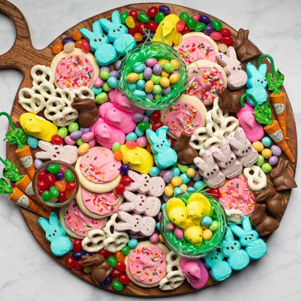 A round wooden board is completely covered in Easter sweets, including pink frosted sugar cookies with sprinkles, blue, pink, and yellow marshmallow Peeps, and small chocolate bunnies. The display is filled in with white yogurt-covered pretzels, green edible grass nests holding candy eggs, and a scattering of vibrant jelly beans.
