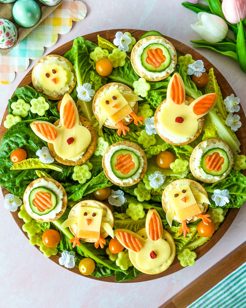 A round wooden board holds several crackers decorated to look like Easter animals resting on a bed of green romaine lettuce. Some crackers feature yellow cheese bunnies with carrot-slice ears, while others show square cheese chicks with carrot beaks and feet. Flower-shaped cucumber slices, cherry tomatoes, and small white flowers are scattered throughout the festive arrangement.