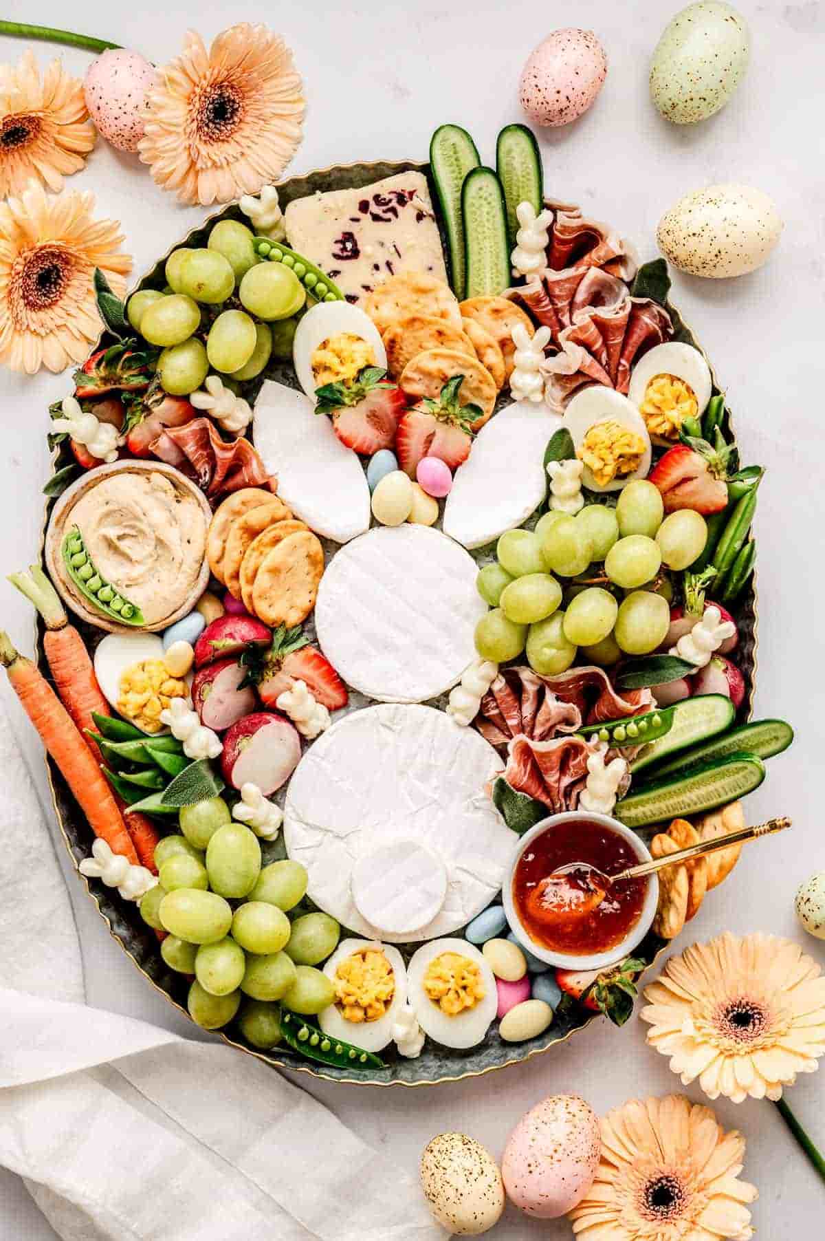 A metal tray filled with an Easter spread centered around rounds of white brie cheese arranged to look like a bunny's head, body, and ears. The board is packed with green grapes, deviled eggs, prosciutto ribbons, cucumbers, snap peas, and radishes. Small white bunny-shaped cheese cutouts, pastel candy eggs, and peach-colored flowers decorate the festive display.
