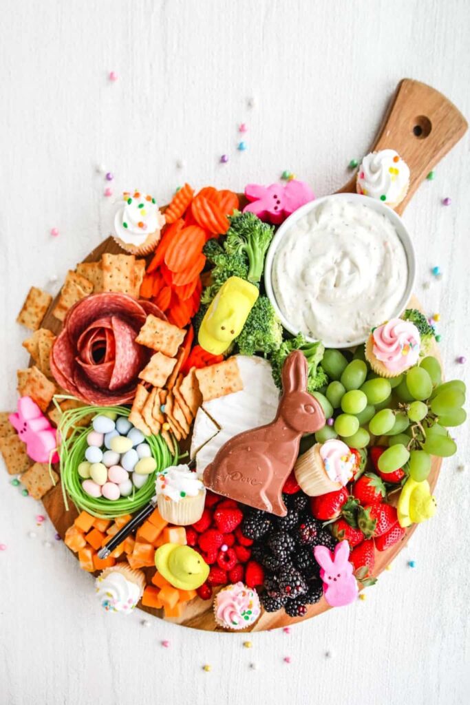 A round wooden grazing board topped with a chocolate bunny, pink and yellow Peeps, and mini cupcakes. It also includes a bowl of white dip, crackers, broccoli, carrots, grapes, strawberries, and a salami rose.