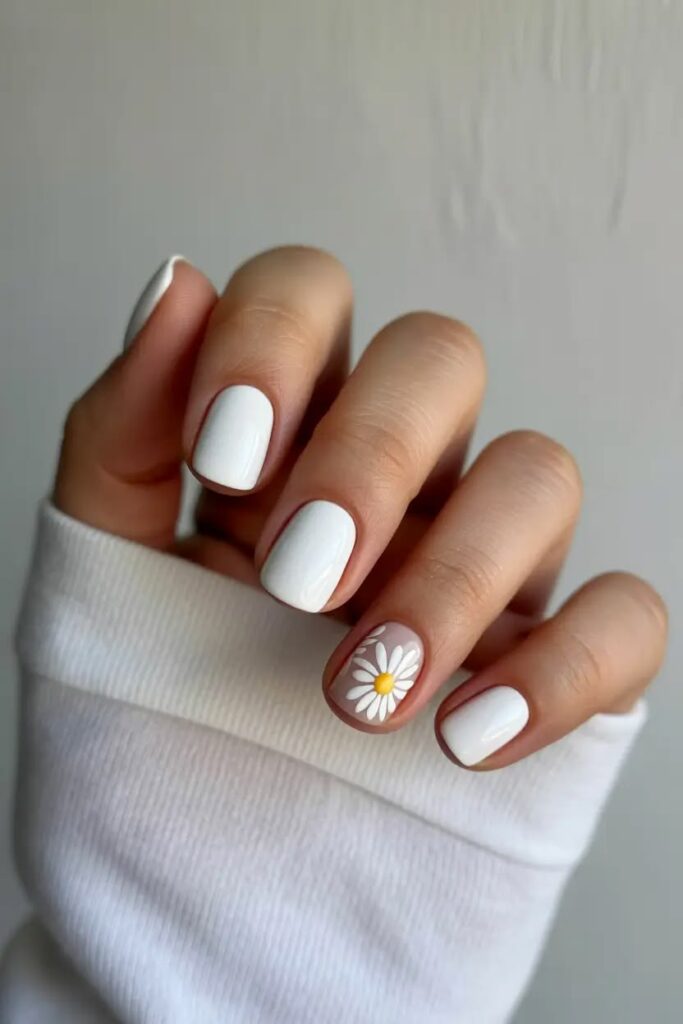 Short square-shaped manicured nails painted in glossy solid white, with a single accent ring finger showing a white and yellow daisy on a clear base.