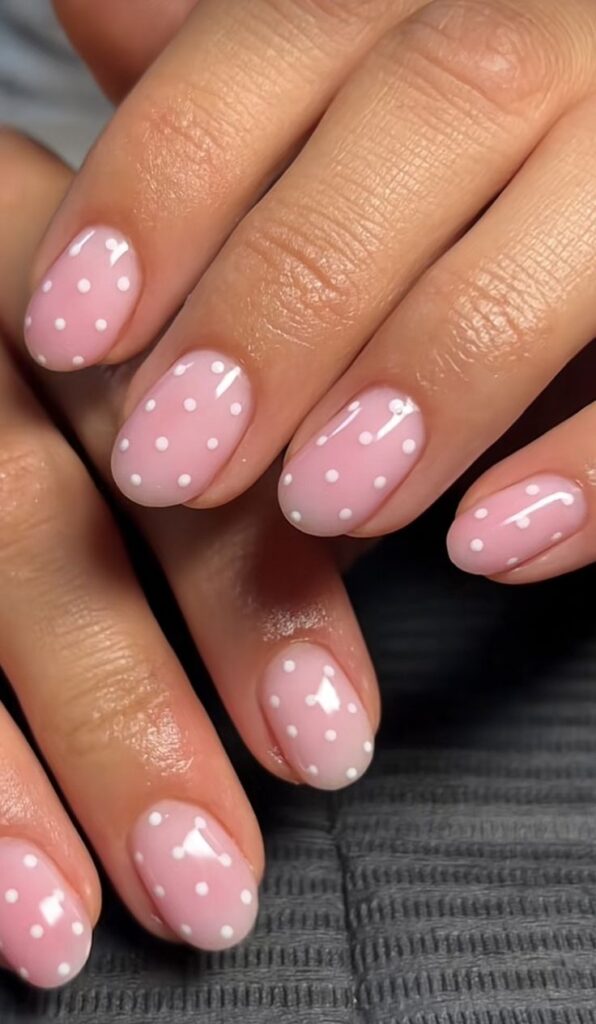 A close-up of round short nails featuring a sheer blush pink base color decorated with tiny white polka dots.