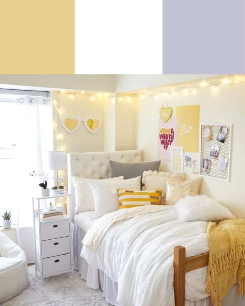 A cozy dorm room featuring a white bed with yellow and grey accent pillows, a yellow throw blanket, white nightstand, and yellow heart-shaped wall decor under warm string lights.