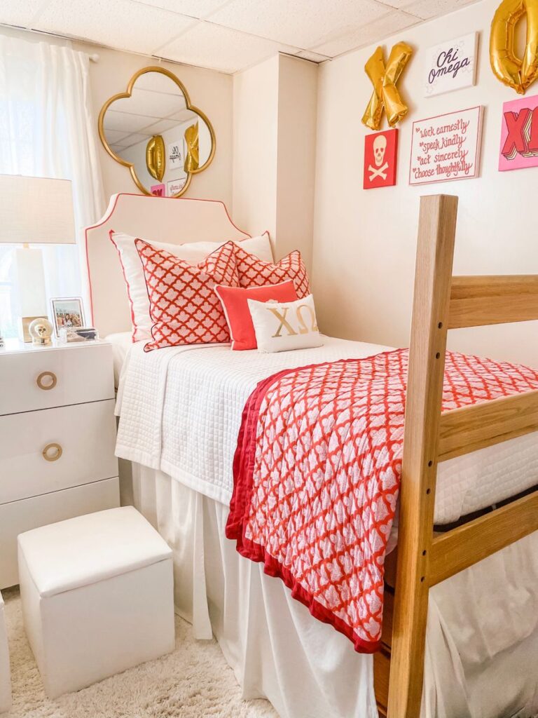 A preppy dorm bed featuring white quilted bedding with red patterned pillows and a matching throw, a white headboard with red trim, and gold wall decor including a quatrefoil mirror and balloon letters.