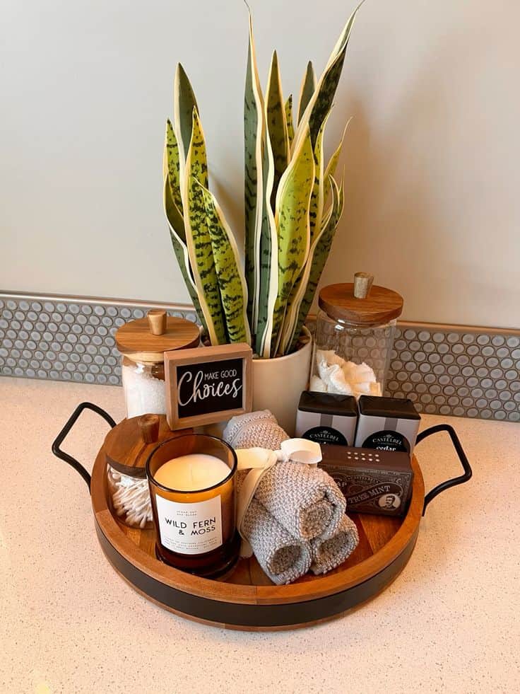 A round wooden tray with black metal handles sits on a white bathroom counter, holding a tall potted snake plant, glass jars with wooden lids, a "Wild Fern & Moss" candle, and rolled grey hand towels tied with a white ribbon.