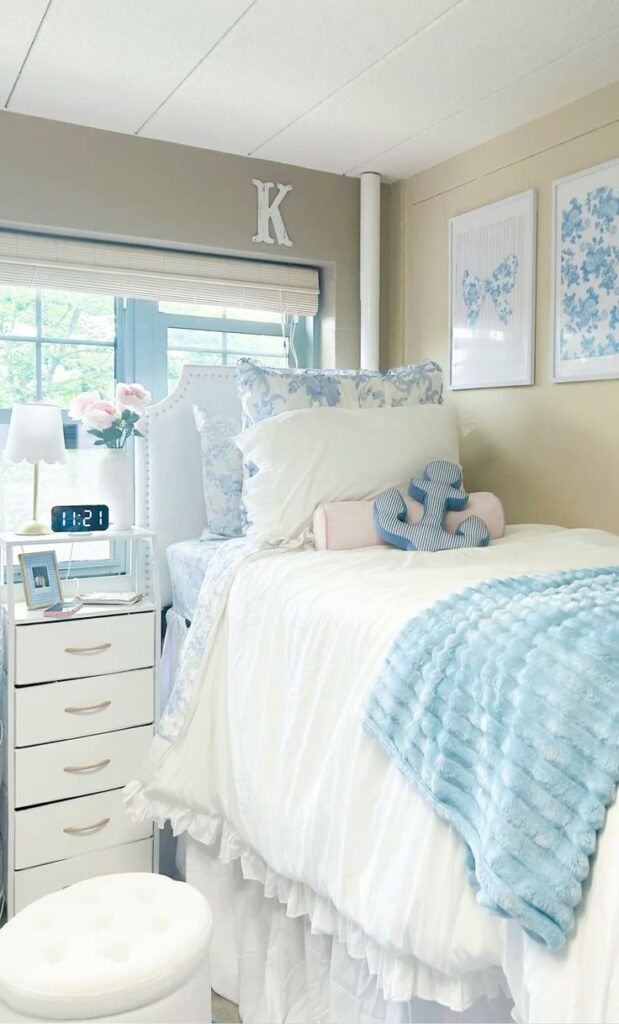 A bright dorm bed with white ruffled bedding, a light blue textured throw, and floral pillows, featuring a white nightstand with a lamp and a large white "K" on the wall above the bed.