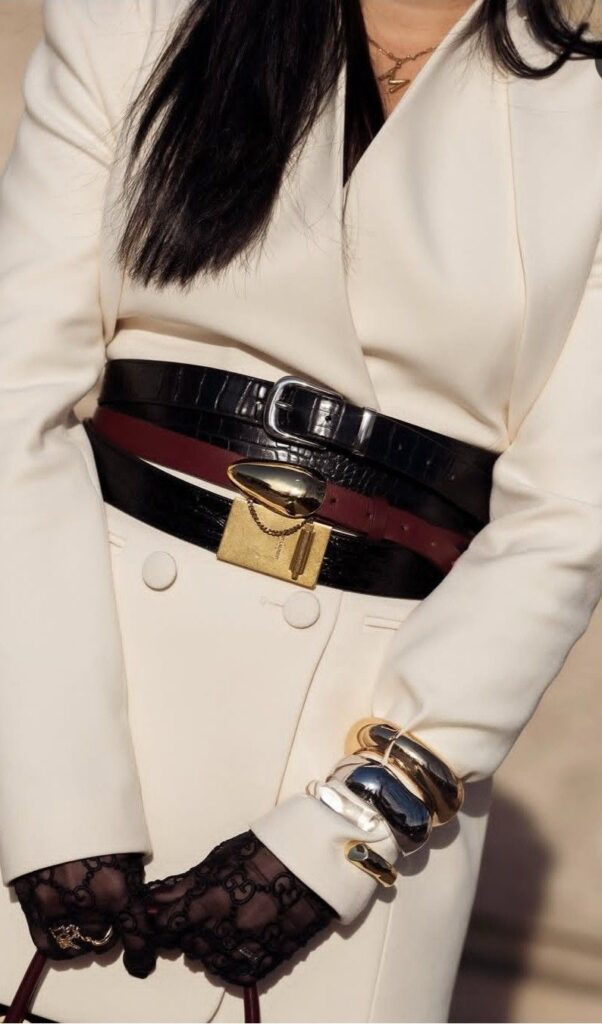 A close-up detail of a white double-breasted blazer cinched with three different layered belts: one black croc-embossed, one thin burgundy, and one black with a large gold buckle. The look includes black lace gloves and chunky gold and silver bangles.