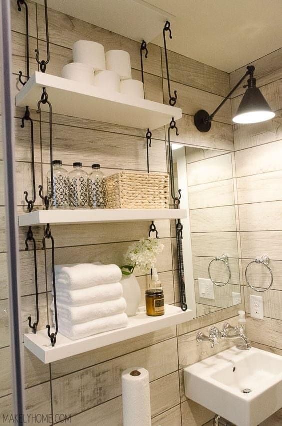 Three white shelves suspended by dark vertical hooks are mounted against a wood-paneled wall, holding toilet paper, wicker baskets, and folded towels.