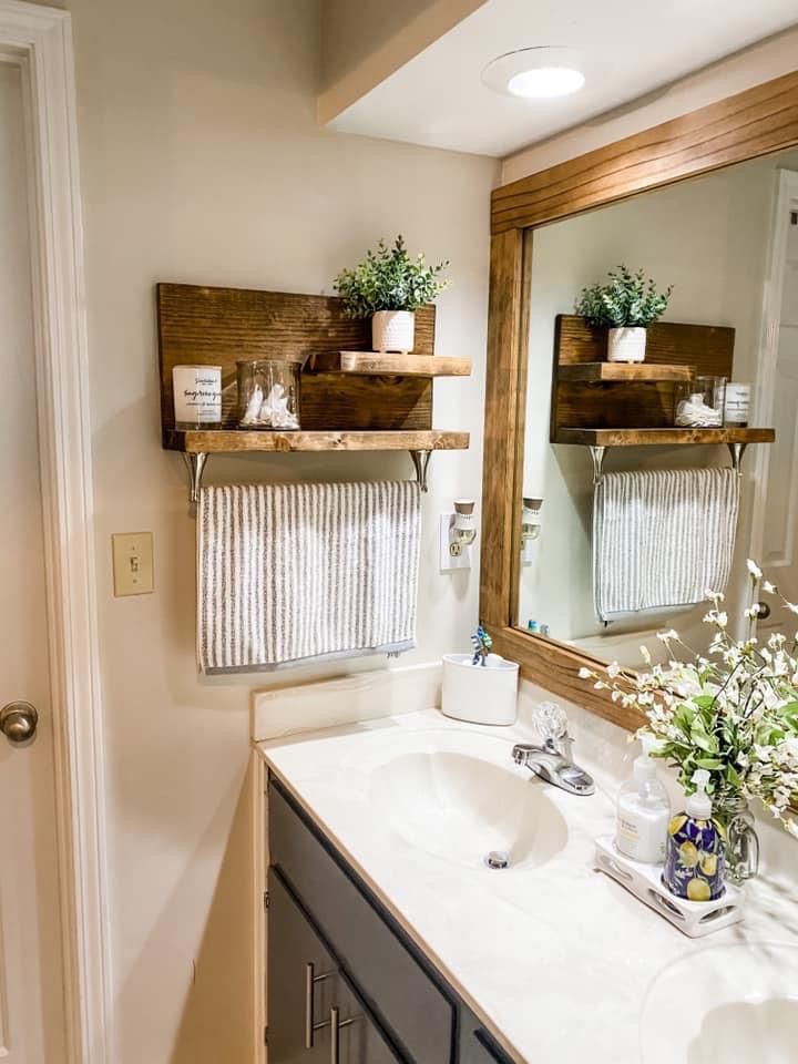 Two levels of wooden shelving are mounted on a beige wall next to a bathroom mirror, with a striped hand towel hanging from a rod on the bottom shelf.