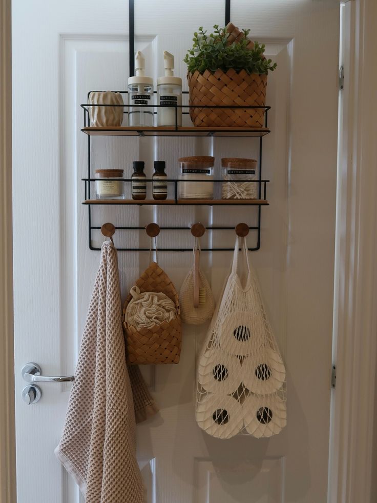 A black metal over-the-door organizer is mounted on a white bathroom door, featuring two wire shelves filled with toiletries and a wooden-peg rack below. A tan towel, a small woven basket, and a mesh bag containing toilet paper rolls hang from the pegs.