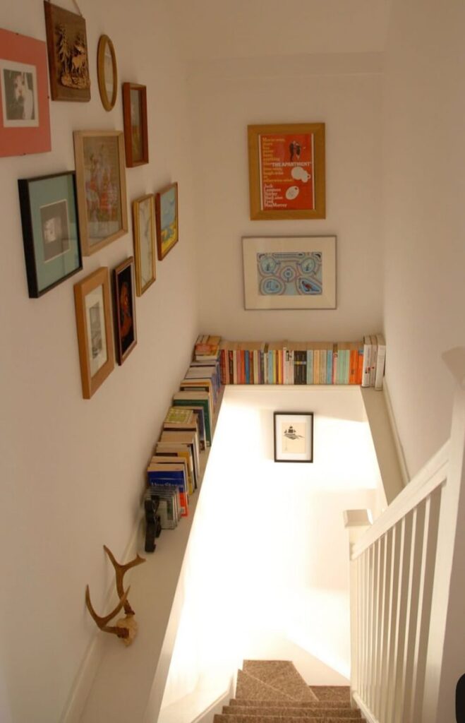 A staircase landing featuring a collection of books arranged along the wall edge, paired with an eclectic gallery wall of framed art prints.