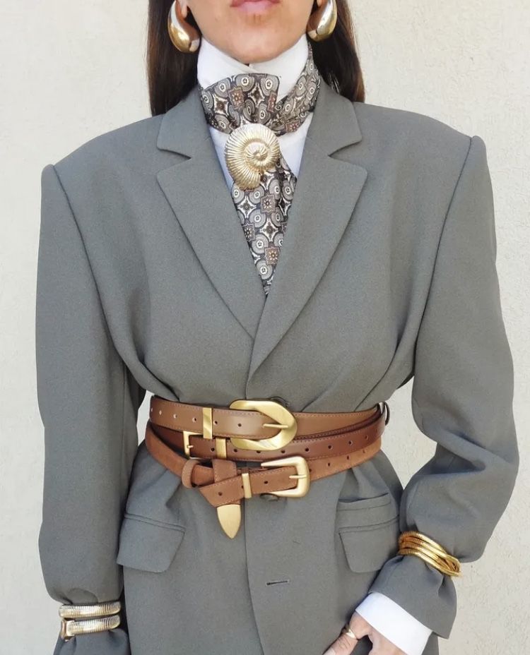 A close-up of a grey oversized blazer cinched with two layered tan leather belts. The outfit is accessorized with a patterned silk neck scarf held by a large gold shell brooch, matching shell earrings, and stacked gold bracelets.
