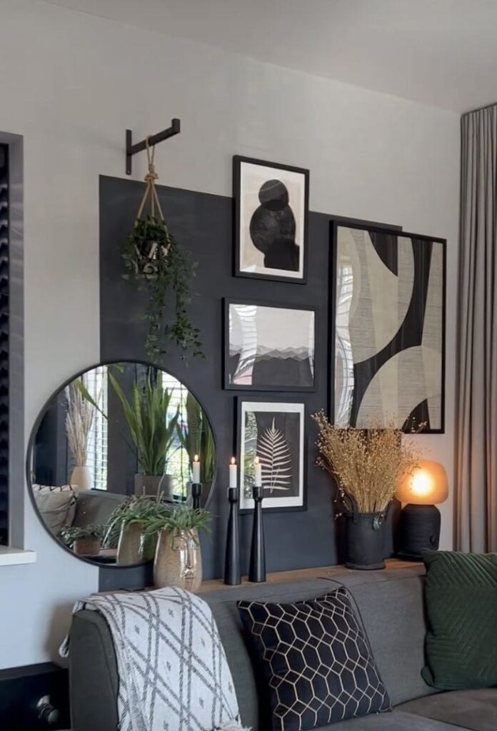 A modern chic living room display behind a dark grey sofa styled with geometric-patterned pillows. The light grey wall features a large, vertically painted charcoal black color-block rectangle that acts as a backdrop for an eclectic gallery. This includes a large round black-framed mirror on the left, a collection of framed abstract and botanical monochrome prints, and a hanging plant suspended from a black wall bracket. A slim wood console table sits behind the sofa, styled with tall black candlesticks, a vase of dried florals, and a small glowing table lamp.