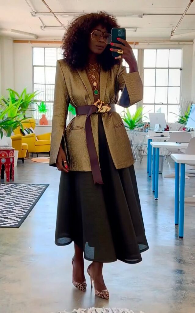 A full-length mirror selfie of a woman wearing a metallic gold textured blazer cinched with a wide burgundy leather belt. She is wearing a long black sheer skirt, leopard-print heels, and oversized gold-rimmed glasses.