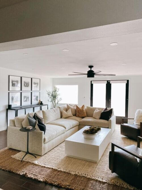 A bright, modern living room featuring a large cream-colored sectional sofa floated away from the wall. Behind the sofa sits a sleek black console table holding a symmetric three-by-two grid of black-and-white framed photos on a white wall. In front of the couch is a chunky woven jute rug layered on dark floors, a solid white rectangular block coffee table, and two dark leather accent chairs. Large black-trimmed French doors and an olive tree in the corner bring natural light and warmth into the minimalist space.