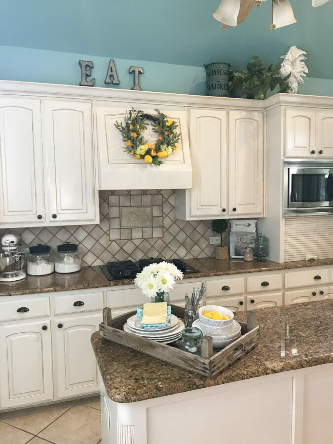 Off-white kitchen cabinets below a bright blue ceiling decorated with metal EAT letters, a faux lemon wreath, and a white rooster figure.