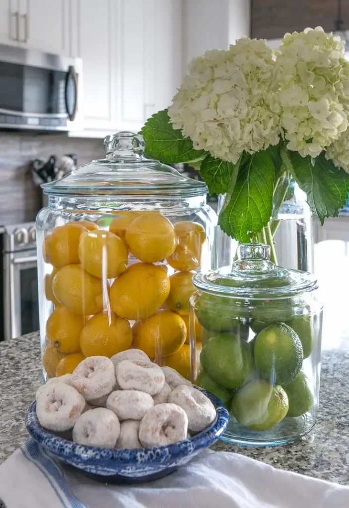 A kitchen counter styled with large glass apothecary jars filled with stacked yellow lemons and green limes, positioned next to a vase of white hydrangeas and a blue bowl of powdered donuts.