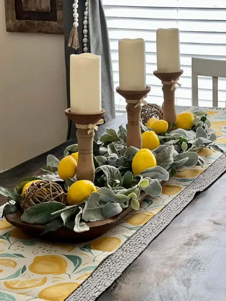 A rustic dining table centerpiece featuring a long wooden dough bowl filled with soft green lamb's ear foliage, loose yellow lemons, twine spheres, and three wooden pillar candlesticks.