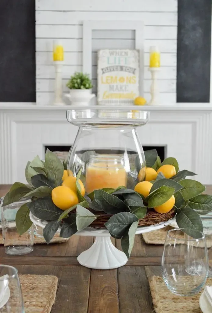 A summer table display featuring a white ceramic cake stand holding a large clear glass hurricane vase with a yellow candle, wrapped in a small grapevine wreath and faux lemon branches.