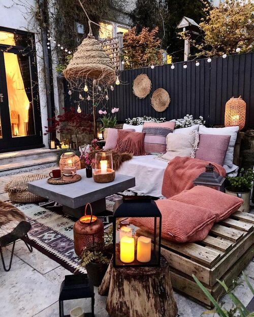A cozy bohemian-style patio at dusk featuring low wooden seating piled with pink and neutral cushions, a grey concrete center table with candles, a large woven pendant light, and string lights along a dark wooden fence.