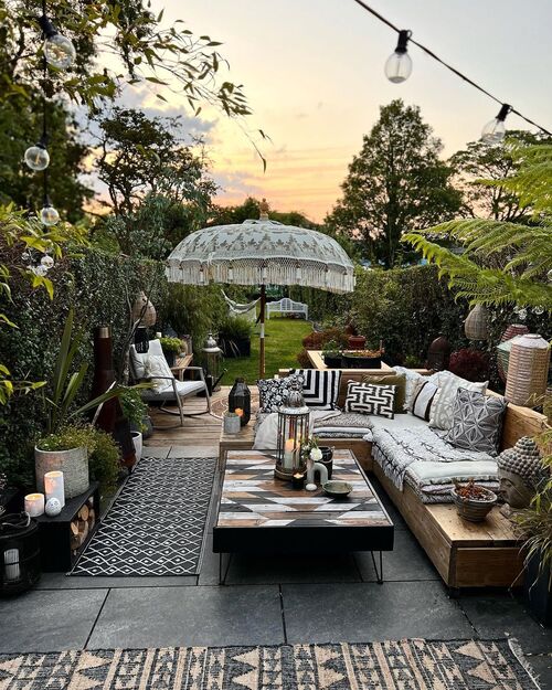 A richly styled backyard patio featuring a wooden L-shaped sectional filled with black and white patterned pillows, a geometric wooden coffee table, a fringed Balinese-style umbrella, and string lights overhead.