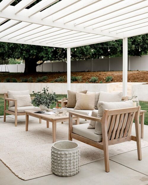 modern patio under a white pergola featuring light wood armchairs and a sofa with cream cushions arranged around a matching coffee table on a large, textured beige area rug.