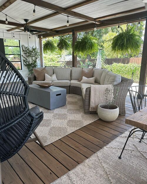 A covered wooden deck featuring a curved wicker sectional sofa, a gray rectangular fire table, a patterned rug, hanging ferns, and string lights illuminating the ceiling.