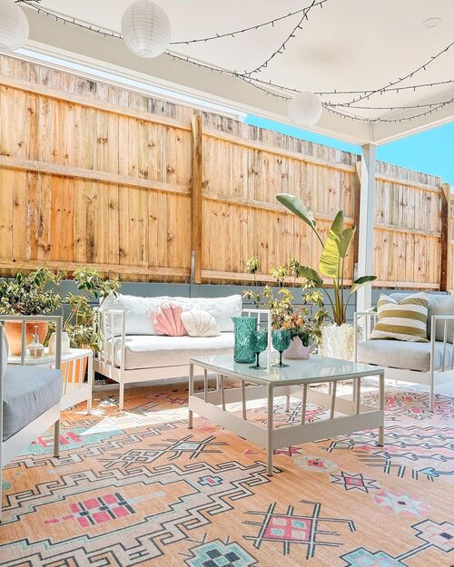 A bright patio under a white roof featuring a colorful peach and turquoise geometric rug, white metal furniture with pastel cushions, potted plants, and paper lanterns hanging from string lights.