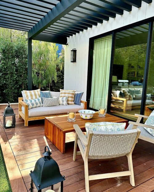 A dark wood deck under a modern pergola featuring a light wood sofa and armchair with striped pillows, a matching wooden coffee table, and large black metal lanterns placed on the floor.