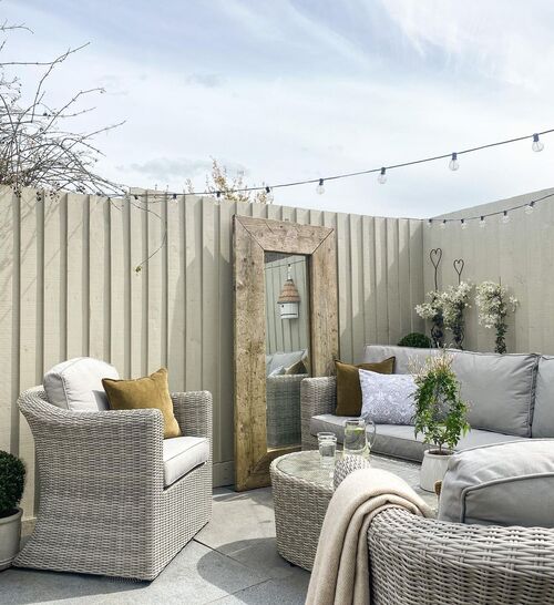 A bright outdoor patio featuring light gray rattan furniture, including a sofa and armchair with neutral cushions, a large vintage-style mirror, and string lights against a white fence.