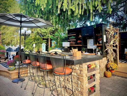 Alt Text: A vibrant, fall-themed outdoor kitchen and bar with stone construction, concrete counters, black-framed shelving, and metal barstools set under evergreen trees.