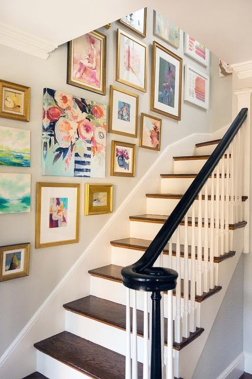 An eclectic staircase gallery wall featuring an assortment of gold-framed abstract prints and colorful floral canvas art hung in a stepped layout along a neutral hallway wall.