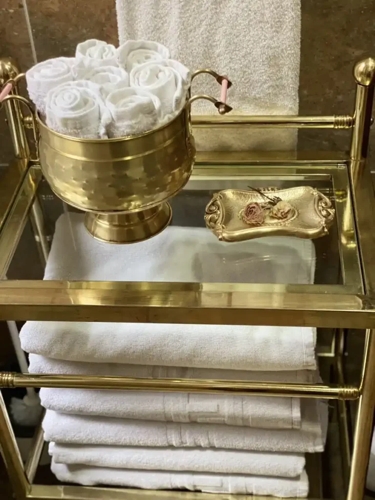 A close-up view of a gold-framed glass shelf unit holding stacked white bath towels. The top shelf holds a hammered gold bowl filled with rolled washcloths and a small gold trinket dish.