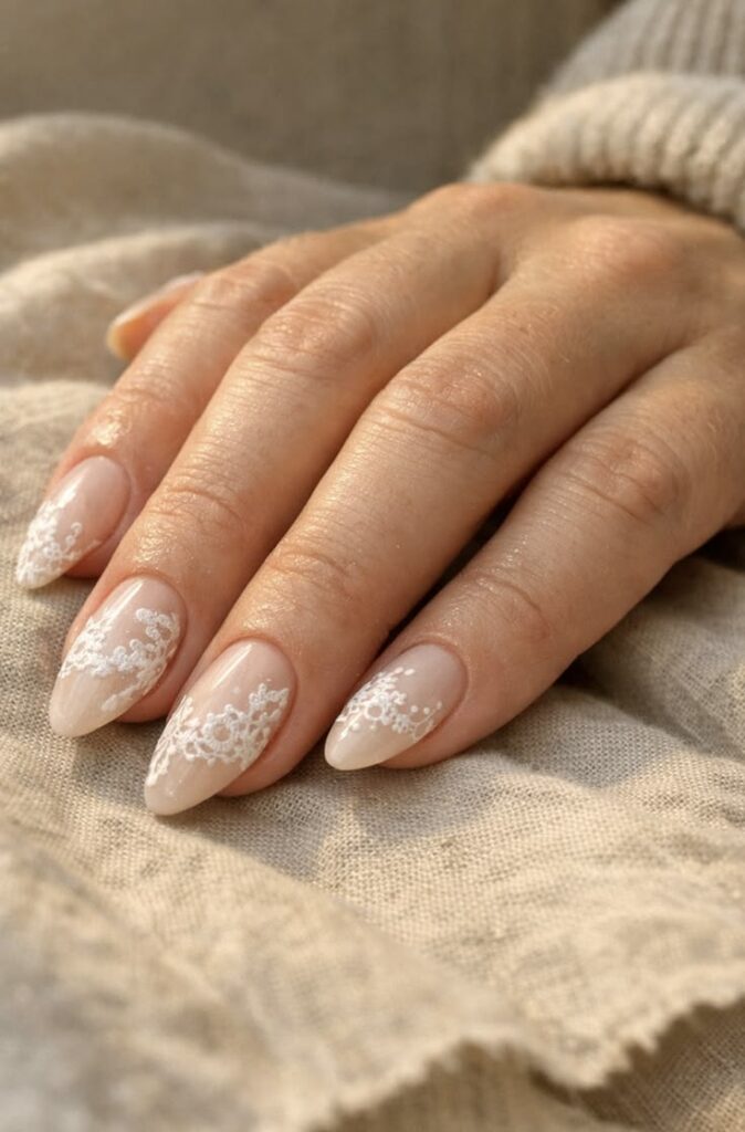 A close-up shot of a hand resting on textured beige fabric, showcasing almond-shaped nails with a sheer nude base and intricate white lace gel nail art painted on the tips.