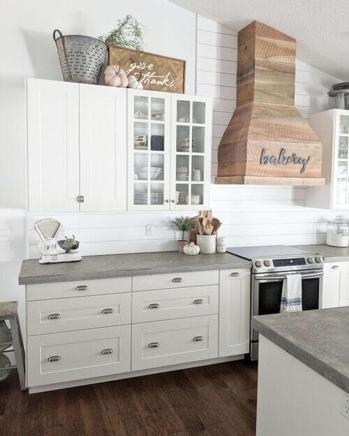 White kitchen cabinets styled for autumn with a large olive bucket, neutral faux pumpkins, and a wooden "give thanks" sign next to a wooden range hood.