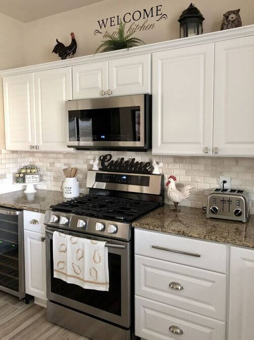 A row of white upper kitchen cabinets decorated with a black rooster, a green fern plant, a black wall vinyl phrase, and a black lantern.