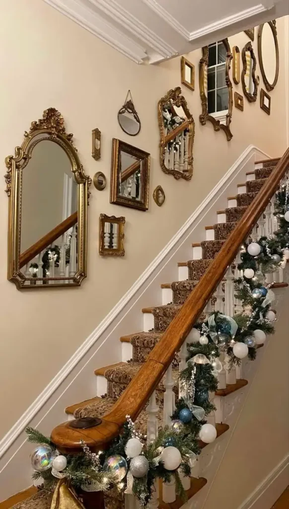 An eclectic mix of vintage ornate gold mirrors arranged in a gallery wall layout along a beige staircase wall decorated with Christmas garland.