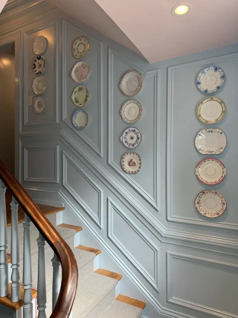 Decorative vintage porcelain plates hung vertically inside light blue decorative staircase paneling and wainscoting molding along a stairway wall.