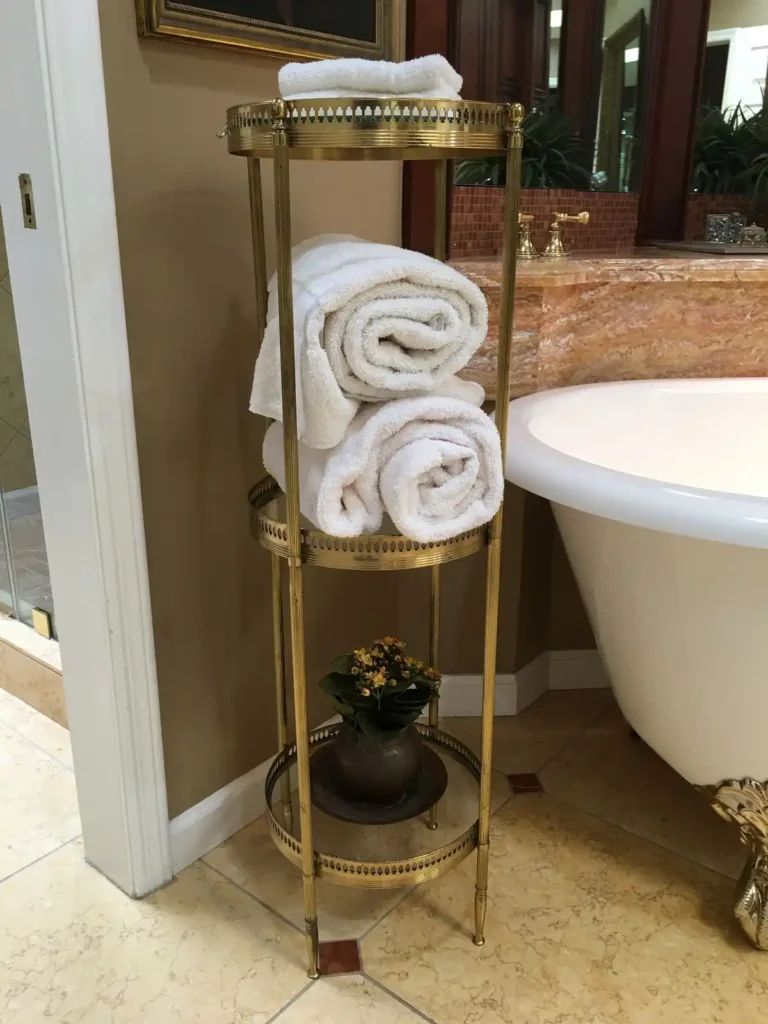 A three-tier round brass towel storage stand stands next to a white clawfoot bathtub. Large rolled white towels rest on the middle shelf, a smaller folded towel sits on top, and a potted plant sits on the bottom glass shelf.