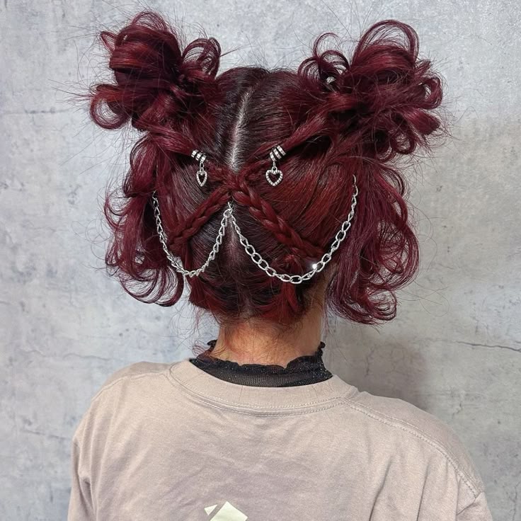 Woman from behind in a tan t-shirt with messy, deep red space buns separated by a middle part that has braids, metal beads, and a hanging chain-and-heart detail, illustrating an edgy grunge prom hair look.