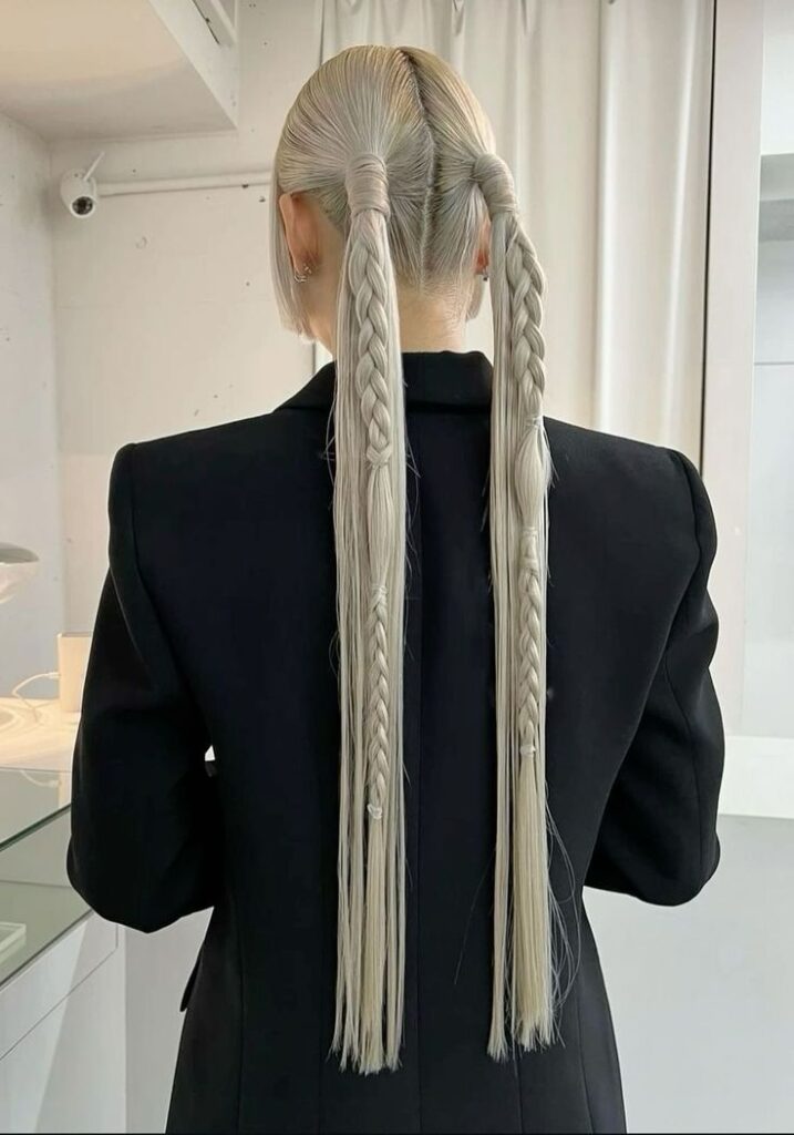 View of a woman from behind in a black blazer, showing her platinum blonde hair in two long, high pigtails that feature thick braids at the top and thin braids with clear bands at the bottom, creating a structured alt prom hair design.