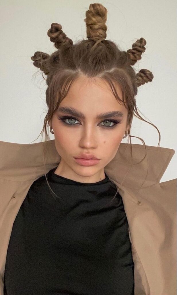 Woman with light brown hair and strong makeup looking at the camera, wearing five small, twisted space buns in an abstract pattern on top of her head, showcasing a grunge prom hair style.