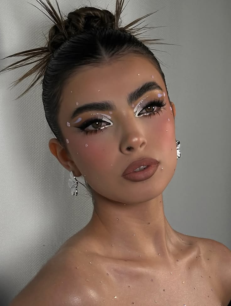 A woman with a grunge prom hairstyle featuring a sleek high bun with spiked ends and white graphic eyeliner with floral accents.
