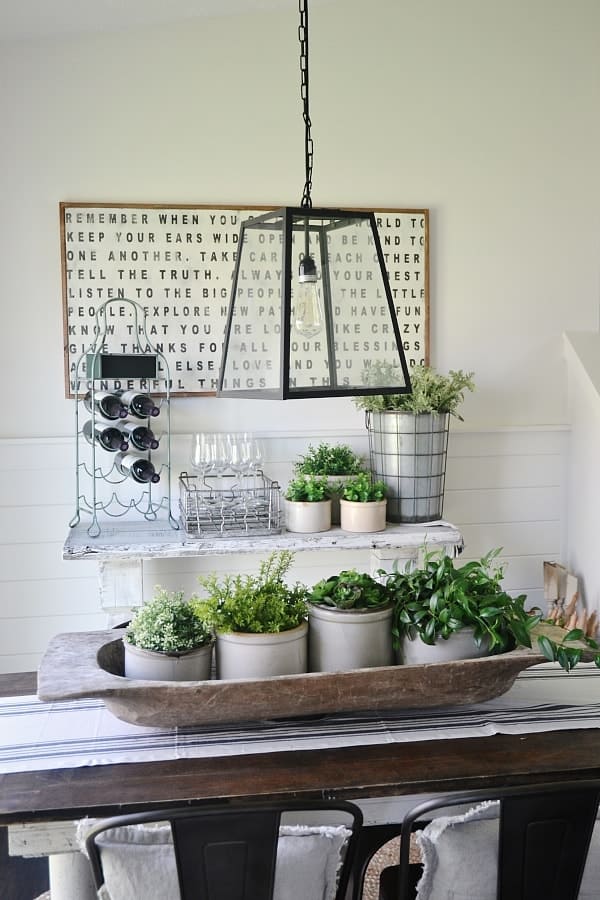 A weathered, oversized summer dough bowl centerpiece sits on a wooden dining table, holding four grey stone crocks overflowing with various green faux plants. This farmhouse decor style offers a lush, organic look that anchors the room's summer table decor.