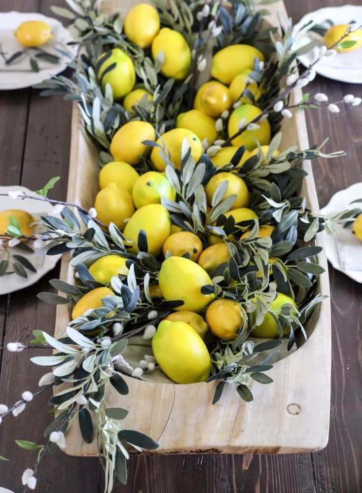 A wooden dough bowl is filled to the top with bright yellow faux lemons and sprays of green olive branches and white pussy willow buds on a dark wood table.