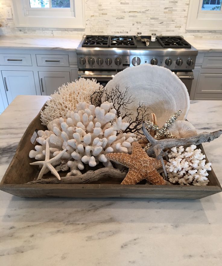 A rustic wooden tray on a marble countertop filled with white coral, dried sea fans, multiple starfish, and a piece of driftwood.