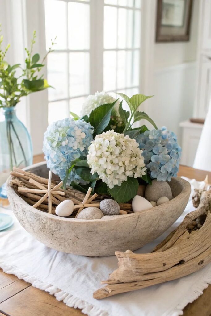 A faux stone bowl filled with white and light blue hydrangea blossoms, green leaves, smooth river stones, and simple stick stars, sitting on a table runner.