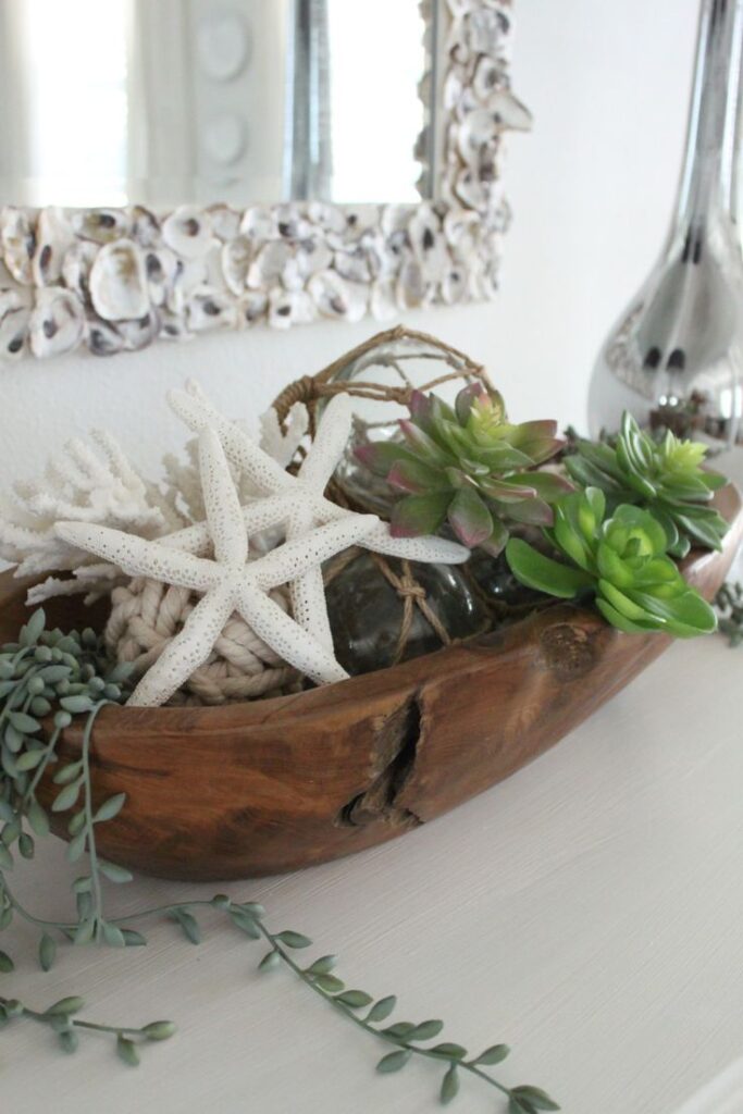A small wooden dough bowl holding white starfish, faux green succulents, coiled rope balls, and a hanging string of pearls plant.