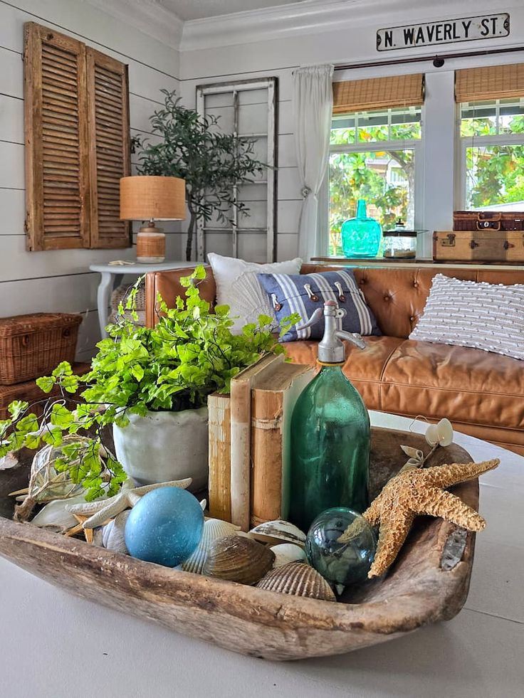  A close-up view of a weathered wooden dough bowl filled with various items like colorful blue and green glass balls, natural seashells, a starfish, two old books, a vintage glass seltzer bottle, and a vibrant green potted maidenhair fern, sitting on a table in a farmhouse style living room.
